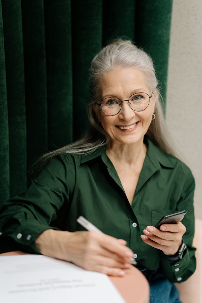 Home Smiling senior woman with gray hair and glasses holding a smartphone indoors.
